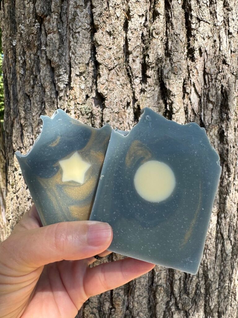 Two Starry Night goat milk soap bars, blue-gray with a cream star and moon, held up against a tree trunk